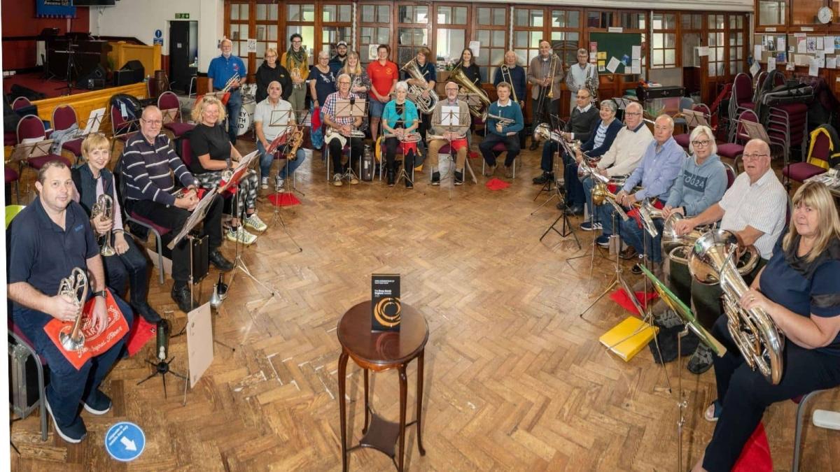 The whole band are sat on chairs in a crescent formation holding their instruments. There is a small wooden table in the middle with a grey and orange awards plaque on it