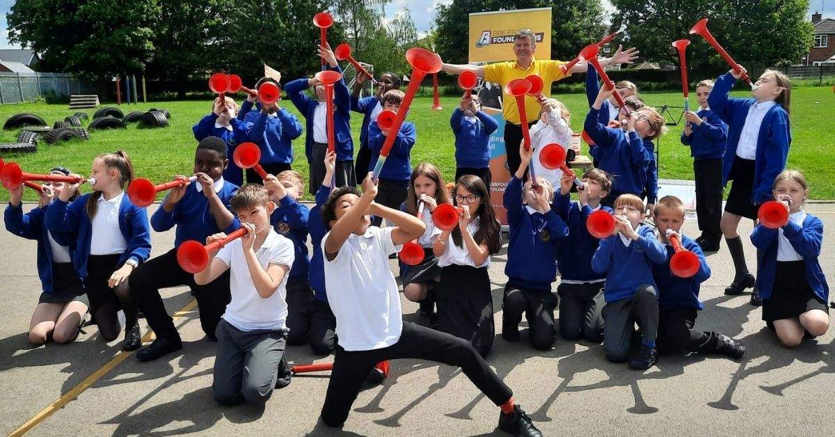 A group of school chilren and their music teacher are standing in front of the school playground whilst each playing a red plastic pBuzz instrument and pointing it outwards or up into the air
