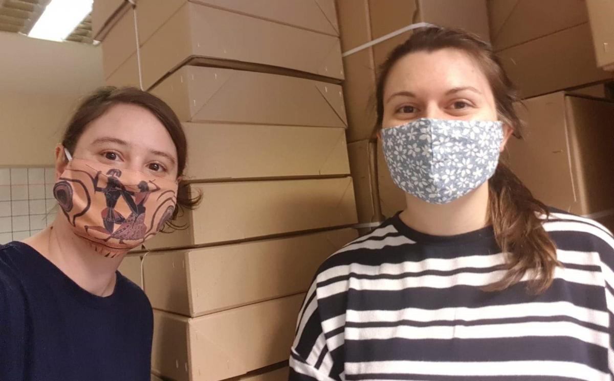 Two women both with brown tied-up hair and a mask standing in front of a tall stack of brown boxes