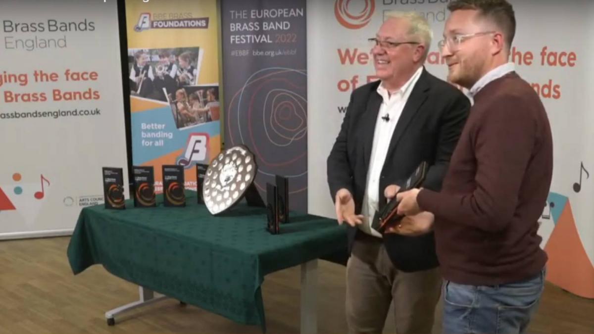 John is posing for a photo with BBE chairman Mike Kilroy, next to a table of award plaques and a large shield