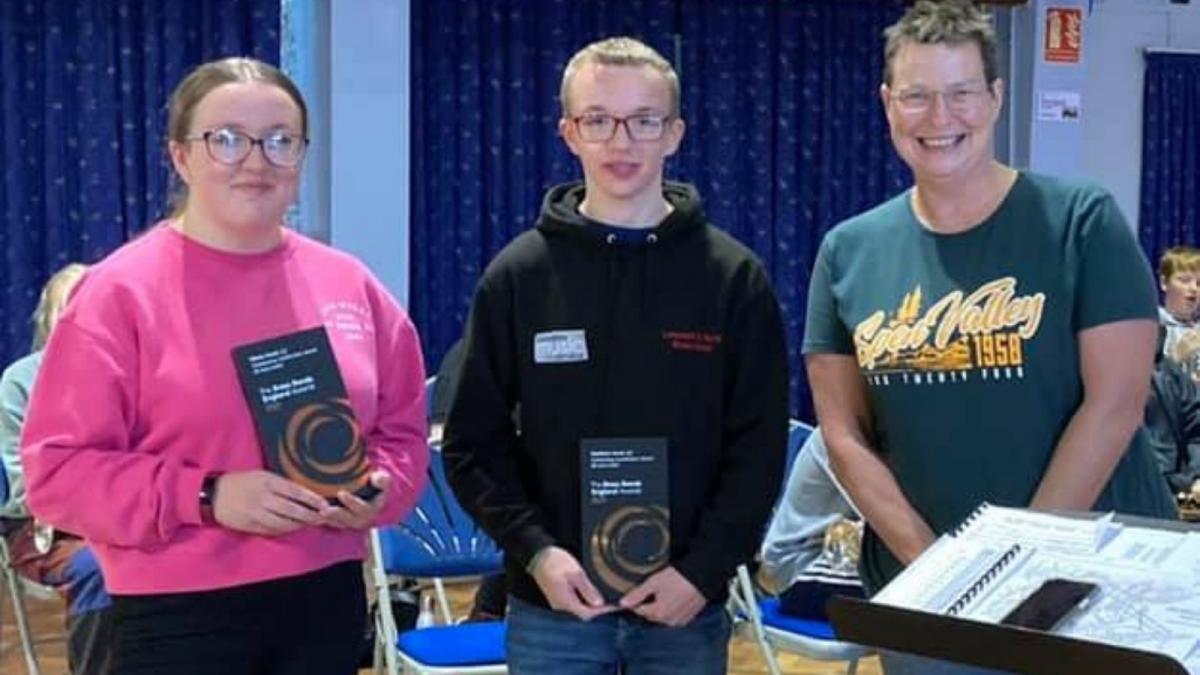 Olivia and Matthew are standing next to their band leader Helen Minshall, both holding a rectangular grey and orange awards plaque