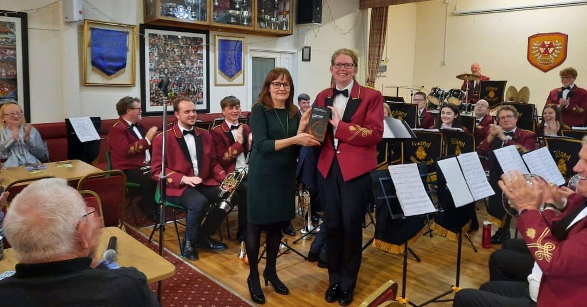 Samantha is posing for a photo with BBE's Rosie Banham, holding her award. They are standing in the middle of the band in the bandroom, are all wearing red band jackets.