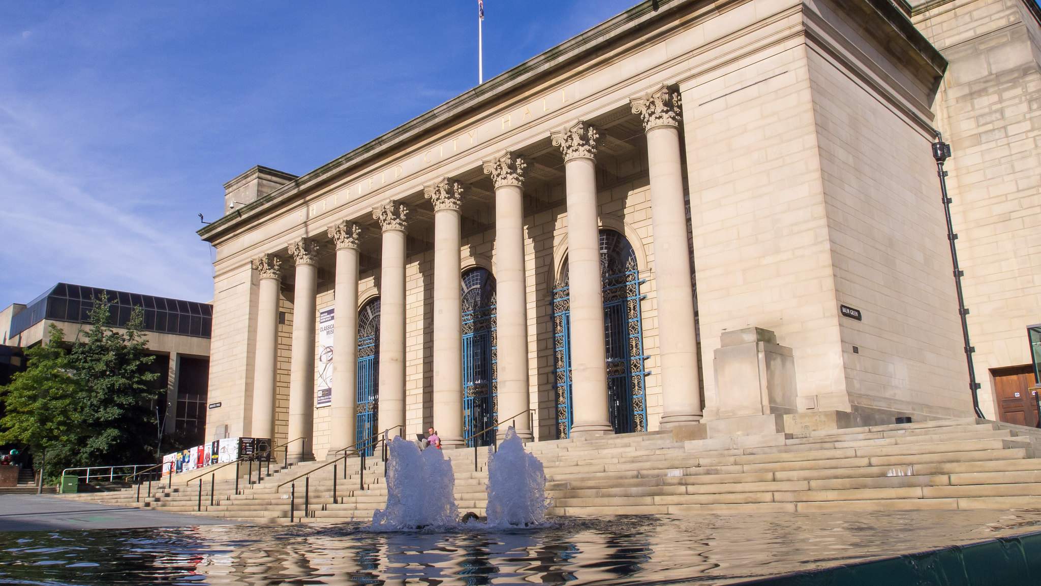 Large building with classical-style collumns and tall windows, behind a pool of water with two fountains in the middle