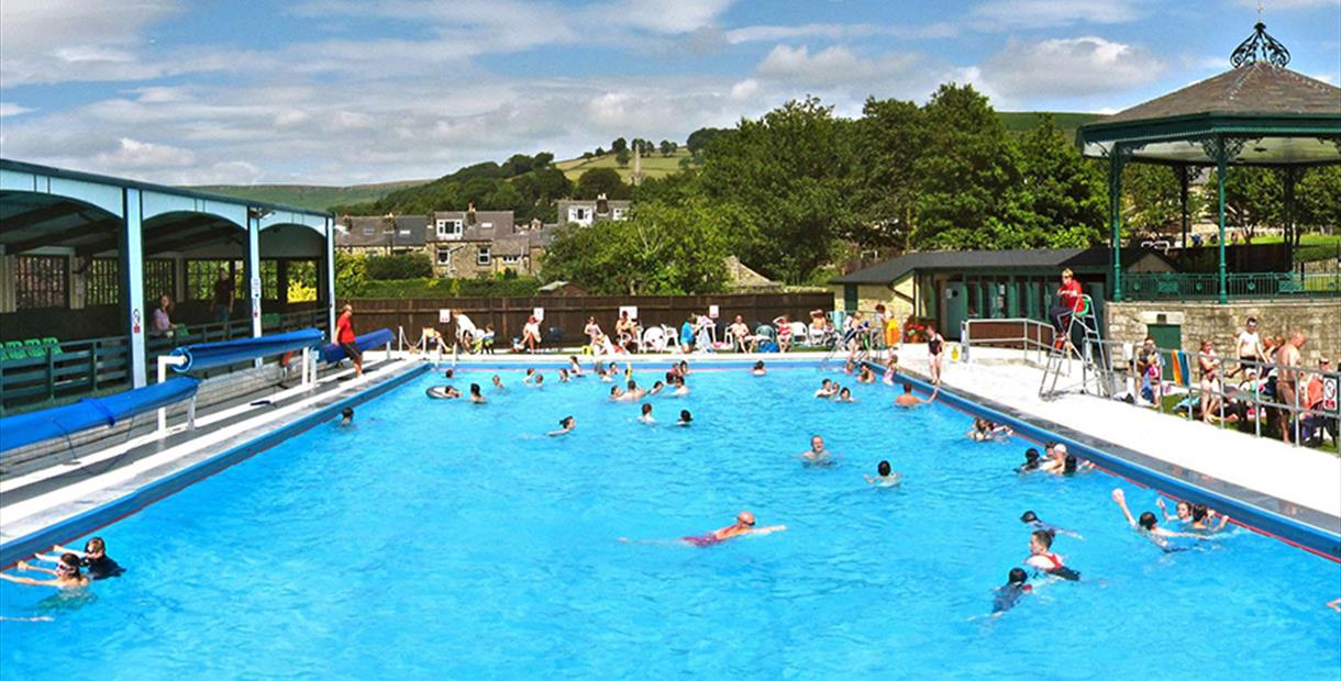 Large outdoor swimming pool with many people swimming and some sat on chairs around the side