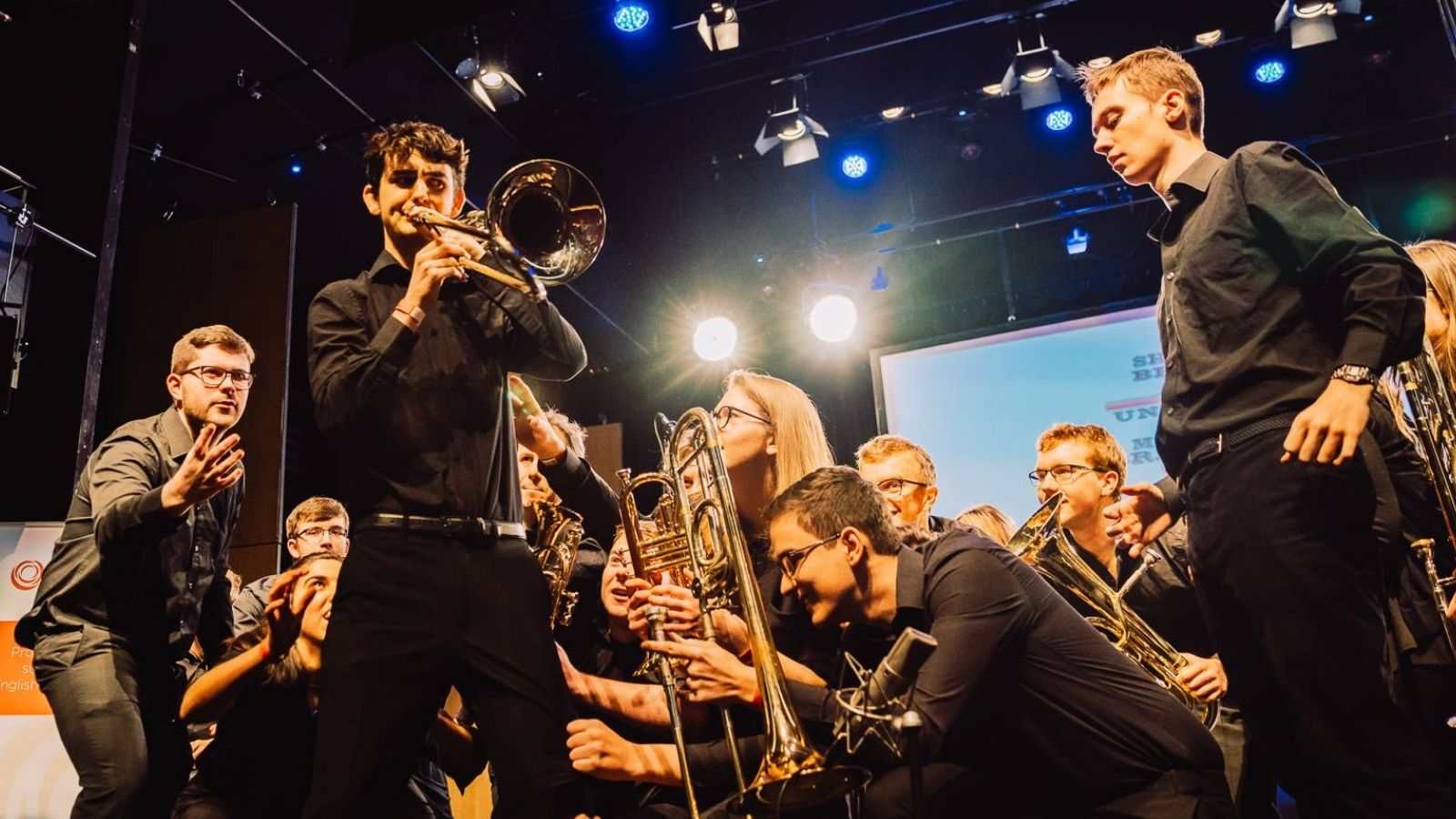 Group of young people holding brass instrumentsm they are all pointing towards a man playing the trombone