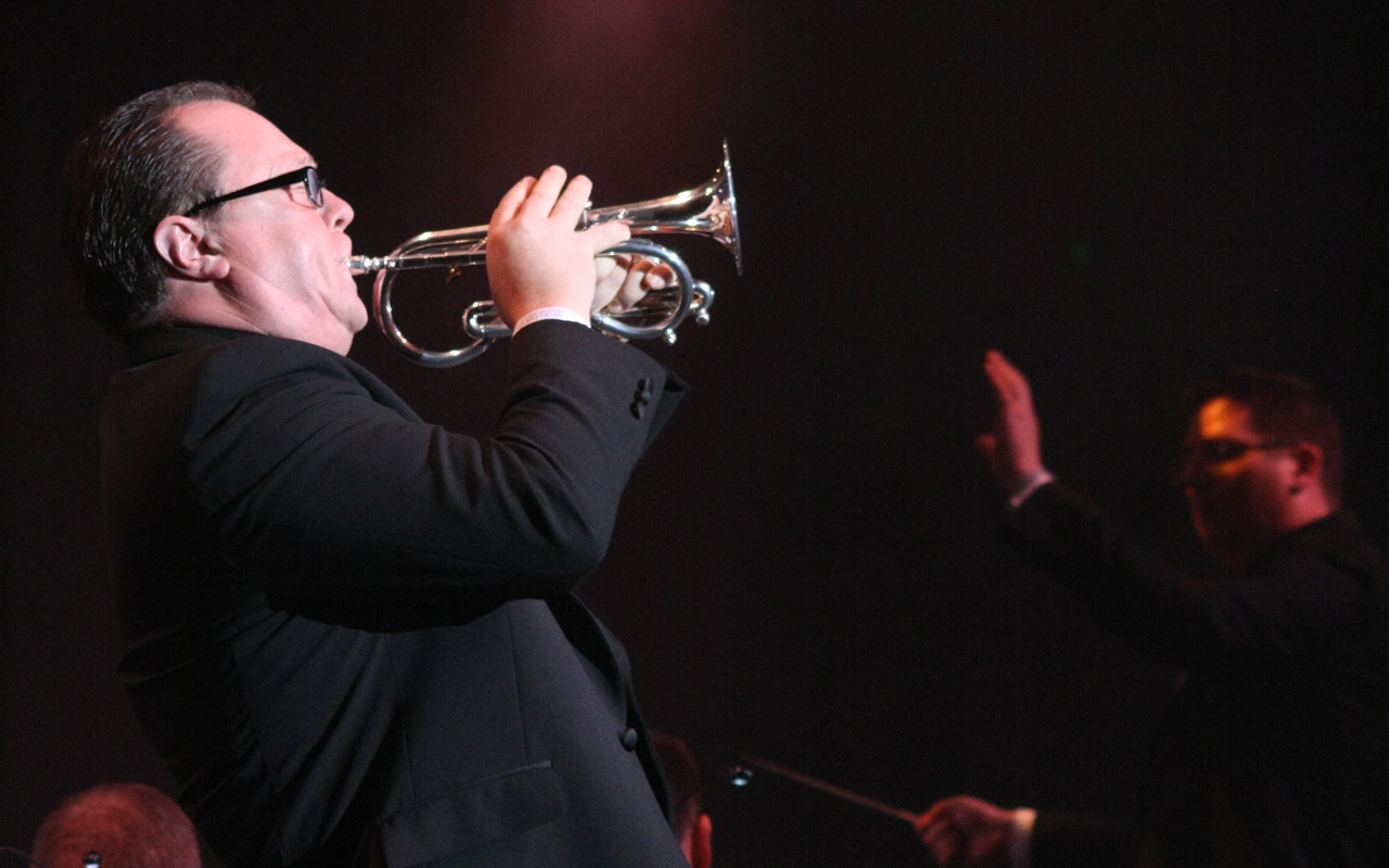 Side-profile of paul playing a soprano cornet. He is wearing a black jacket and glasses. The room is dark and a conductor can be seen in the distance