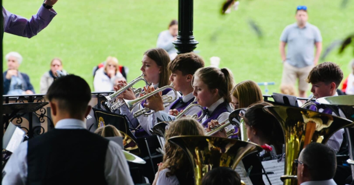 A group of youth brass players performing in a bandstand. An audience can be seen in the background on the grass.