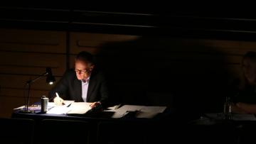 Man wearing glasses sat in dark at long table with lamp, writing on sheets of white paper, bottle of water in front of him