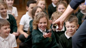 Children wearing green sweatshirts sitting on a wooden floor - a girl reaches forward to take a small brass instrument