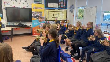 Group of children all facing left each playing a cornet, they are wearing blue sweatshirts with yellow polo shirts