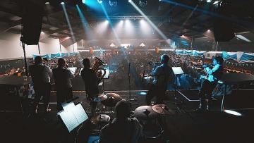 Five brass players and a drummer stand on stage facing a large crowd, inside a venue with spotlights shining on them