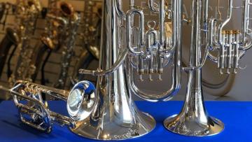 Upturned brass instruments on a table with blue cloth in front of a rack of saxophones