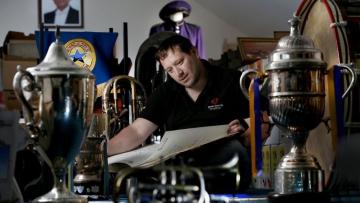Man with brown hair and black polo top is reading a large score surrounded by trophies