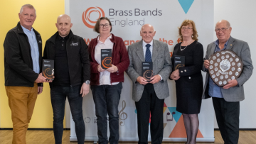 Four men and two women standing in front of a banner reading "Brass Bands England", each holding a grey and orange Award plaque and smiling