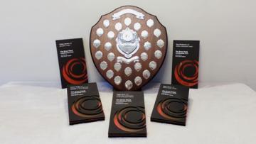 Large wooden shield covered in small engraved metal plaques, surrounded by five rectangular grey and orange awards