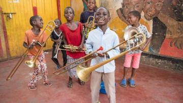 Group of six young boys carrying a trombone or cornet are standing in front of a red floor in front of a red and yellow mural