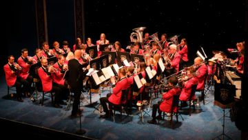 Burbage band members are sitting down in a circle band formation playing instruments, wearing red brass band blazors, the conductor is standing at the front facing the band
