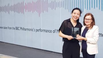 CJ Wu and BBE's Rosie standing in front of a large white backdrop with a soundwave. Rosie is wearing a white jacket and CJ is wearing black. They are both holding a clear rectangular plaque