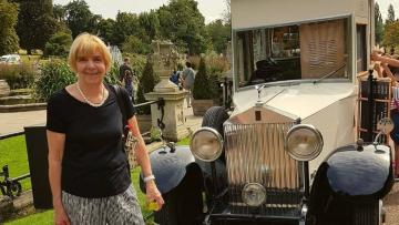 Carole has short blonde hair and is wearing a black t-shirt and black and white patterned trousers. She is standing in a park next to a vintage truck which is serving ice creams.