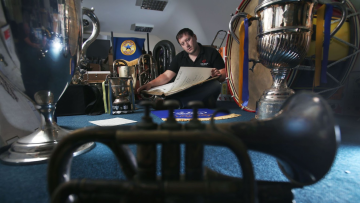 Parky of the BBE Team sits surrounded by items from the Brass Bands Archive. He holds a large book open on his lap