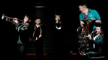 Line of children wearing green polo tops and playing brass instruments in front of a black backdrop