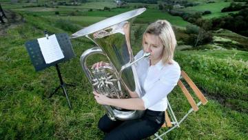 Woman is say on a wooden chair on a grassy hilltop, next to a music stand, she is playing a bass, she has blond hair and is wearing a white shirt and black trousers