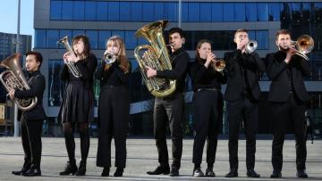 Seven musicians dressed in black and holding brass instruments standing in front of the glass exterior of Symphony Hall Birmingham