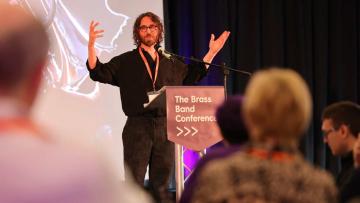 Gavin has curley brown hair and dark glasses, he has his hands outstretched and raised, he is standing behind a lecturn with 'The Brass Band Conference' in bold white lettering on a purple background