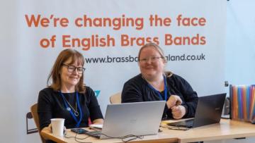 Two women with brown hair and wearing black tops with blue lanyards siting in front of a sign that read's "We're changing the face of English Brass Bands"