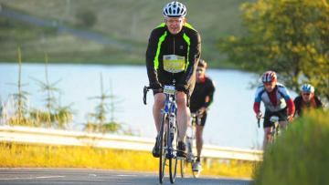 Kenny is cycling on a road in front of a lake. He is earing black and luminous yellow cycling gear and a white helmet