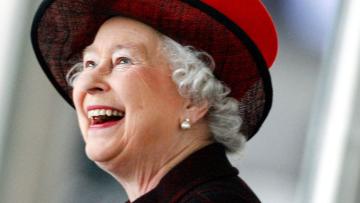 Queen looking to the left and smiling, she has a red hat on and you can see a black coat collar
