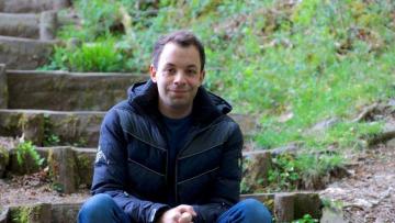 Sam is sitting on the bottom of a series of wooden steps next to a patch of grass he has brown hair and is wearing a navy blue coat and jeans