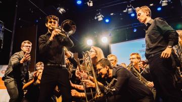 Group of young people holding brass instrumentsm they are all pointing towards a man playing the trombone