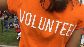 A women's back she is wearing an orange t-shirt which says Volunteer on the back in white all caps