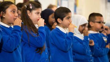 Five children holding different coloured plastic mouthpieces to their mouths. They are wearing blue sweatshirts and white colors