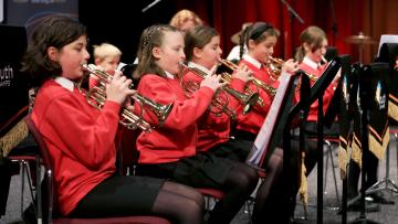 Line of girls playing brass insturments, they are wearing red sweatshirts and black trousers or skirts