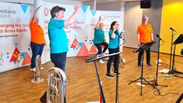 Five musicians each holding a brass instrument and wearing a different brightly coloured t-shirt are waving their arms in the air