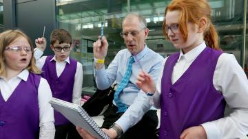 man in blue shirt conducts with a pen, teaching 3 young people in purple waistcoats to do the same