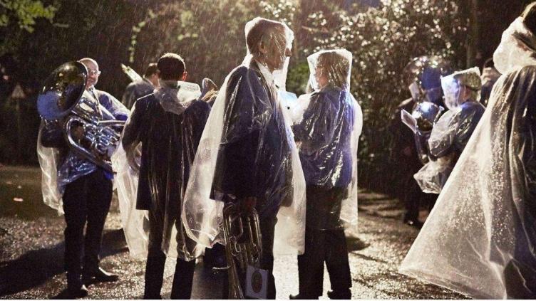 Group of people in rain at dusk are carrying brass instruments and wearing clear rain ponchos