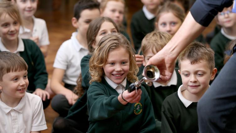 Children wearing green sweatshirts sitting on a wooden floor - a girl reaches forward to take a small brass instrument