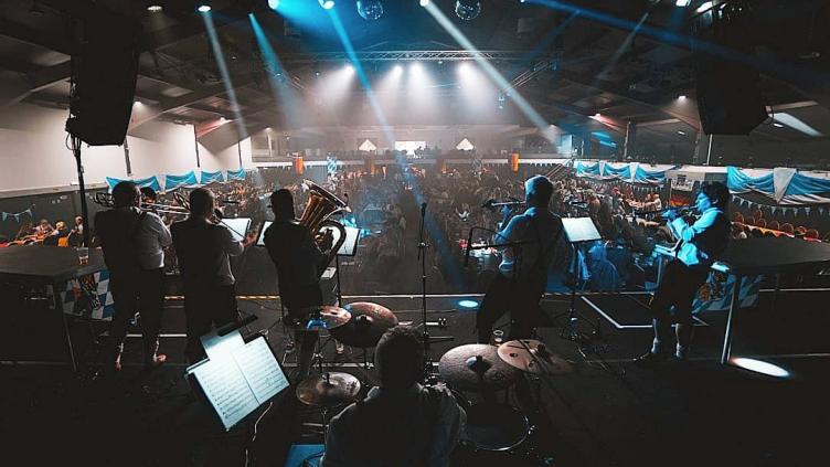 Five brass players and a drummer stand on stage facing a large crowd, inside a venue with spotlights shining on them