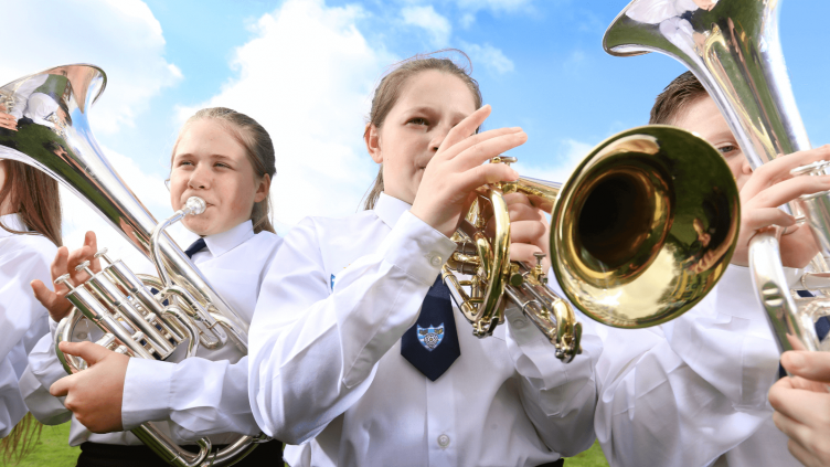 Two girls and a boy playing silver brass instruments in front of a bright but cloudy sky