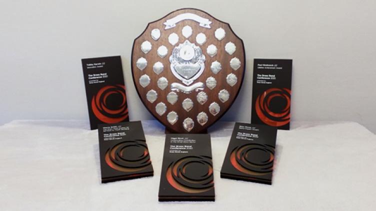 Large wooden shield covered in small engraved metal plaques, surrounded by five rectangular grey and orange awards