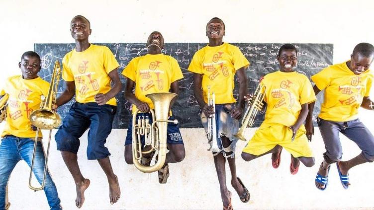 Group of six young people jumping in the air in front of a black board, holding brass instruments. They are wearing bright yellow t-shirts and jeans.