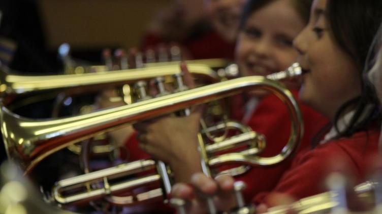 Close up of three children each playing a cornet, they are each wearing a red jumper