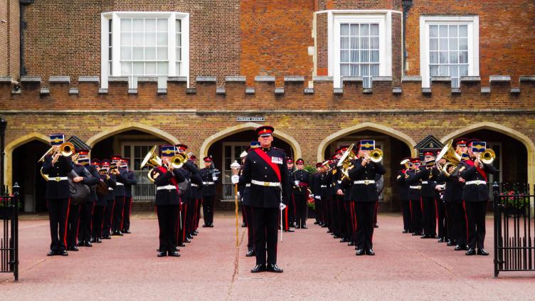 Band members are standing in line on a forecourt in front of a red brick building