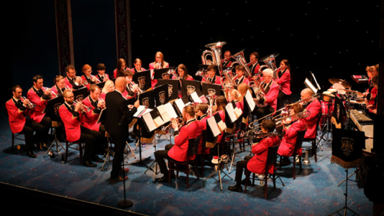 Burbage band members are sitting down in a circle band formation playing instruments, wearing red brass band blazors, the conductor is standing at the front facing the band