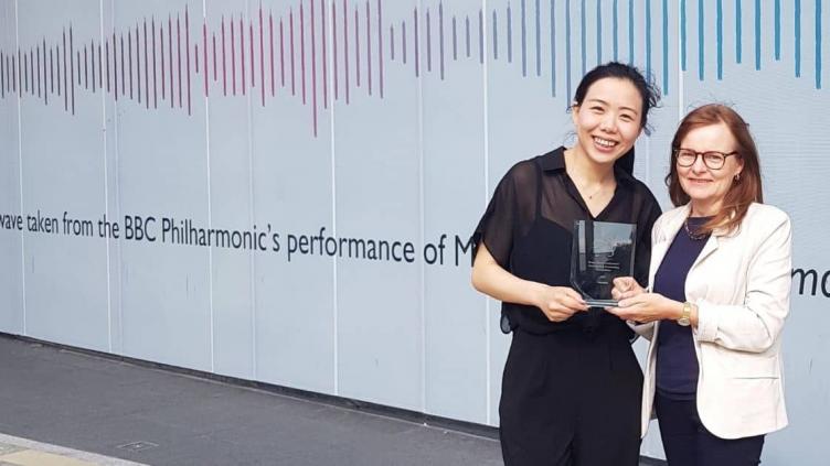 CJ Wu and BBE's Rosie standing in front of a large white backdrop with a soundwave. Rosie is wearing a white jacket and CJ is wearing black. They are both holding a clear rectangular plaque