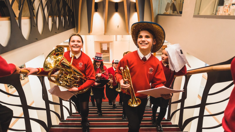 Young brass players walk up a staircase with their instruments. They are smiling and talking. One of them wears a Stetson as a performance prop. 