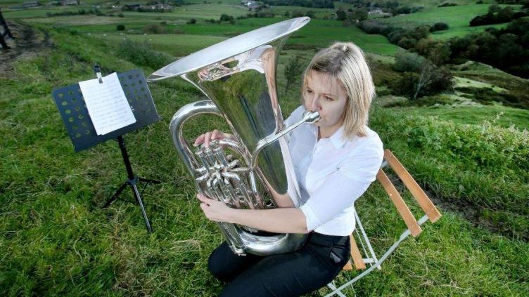 Woman is say on a wooden chair on a grassy hilltop, next to a music stand, she is playing a bass, she has blond hair and is wearing a white shirt and black trousers